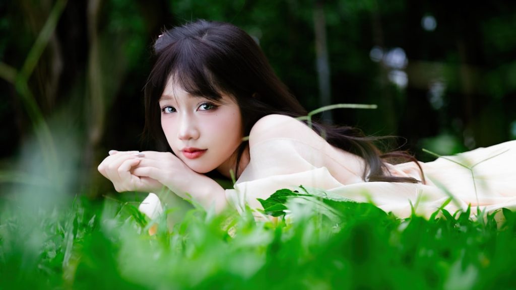 Smiling young woman lying on green grass with a bouquet of fresh hydrangea flowers, wearing a soft cream dress in natural light.