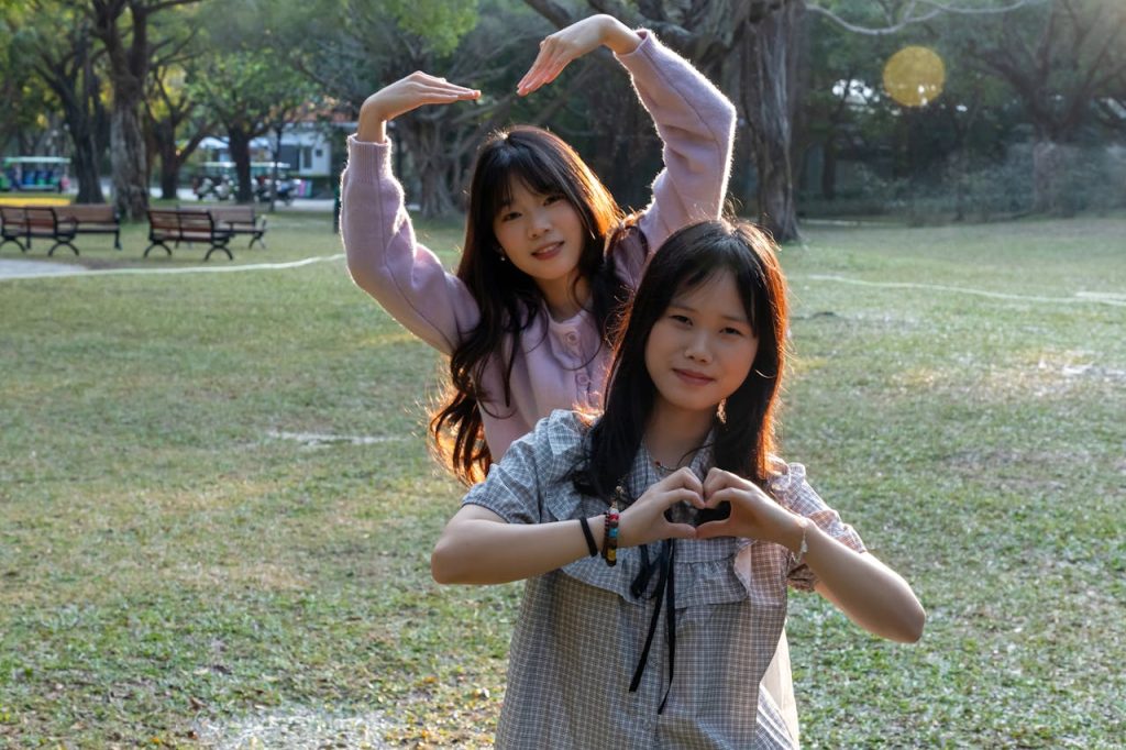 Two young girls making heart shapes with hands in a sunny park setting.