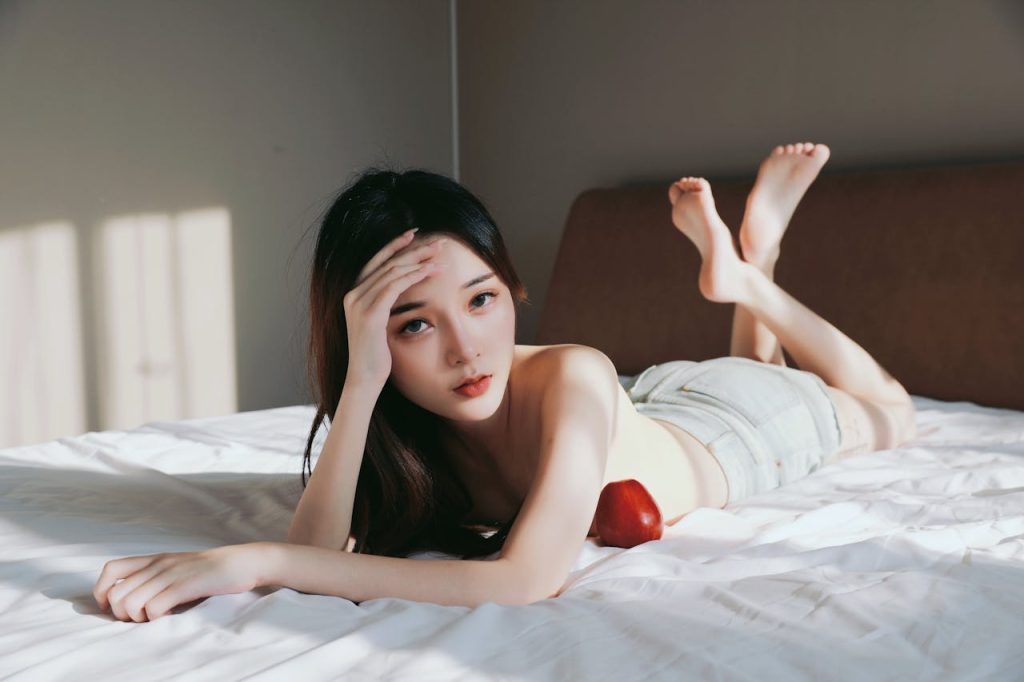 Asian woman relaxing on a bed, barefoot with an apple, in soft natural light.
